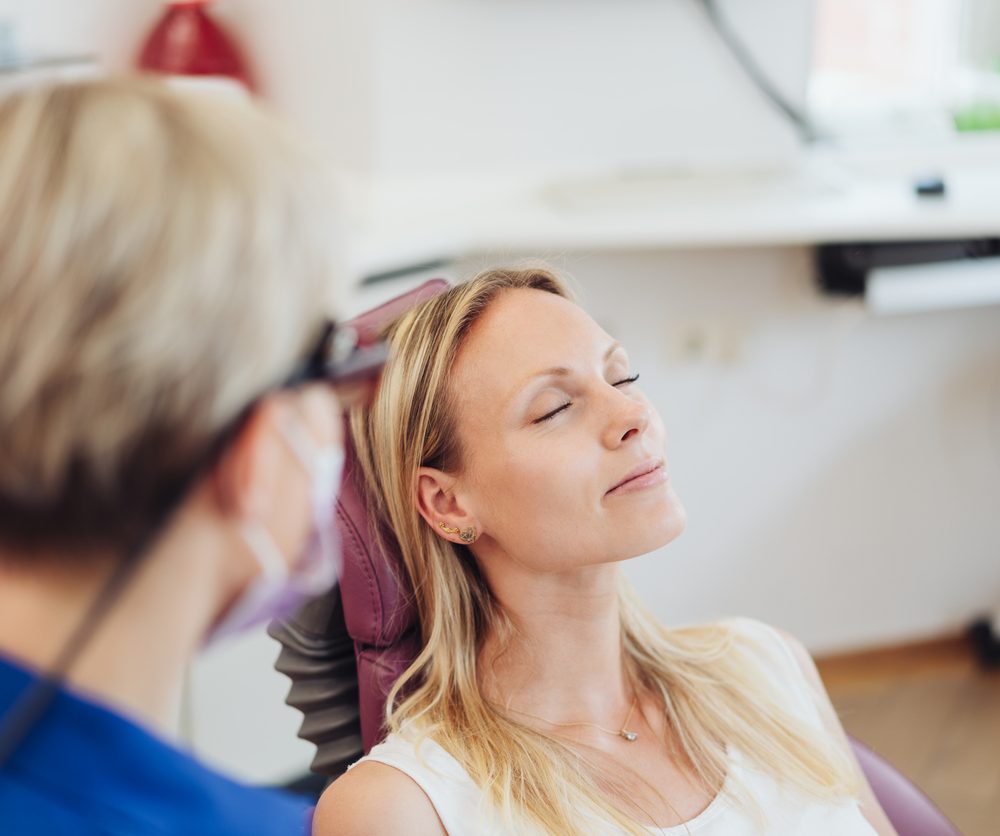 Woman relaxing with her eyes close at the dentist as she sits in the examination chair viewed over the shoulder of the dentist or nurse